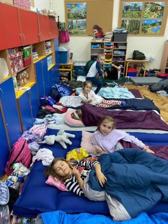 Uczniowie leżą w spiworach i na materacach w sali lekcyjnej, przygotowują się do snu.