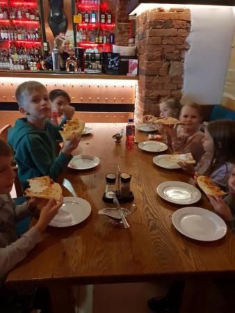 Uczniowie klasy 2b siedzą przy stole w restauracji i jedzą pizzę.