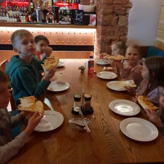 Uczniowie klasy 2b siedzą przy stole w restauracji i jedzą pizzę.