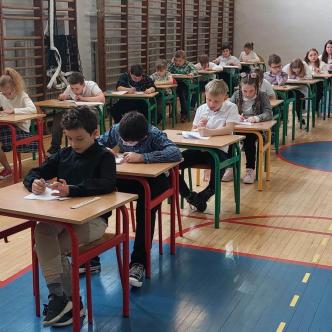 Uczniowie siedzą w ławeczkach na sali gimnastycznej i piszą ogólnopolski test kompetencji